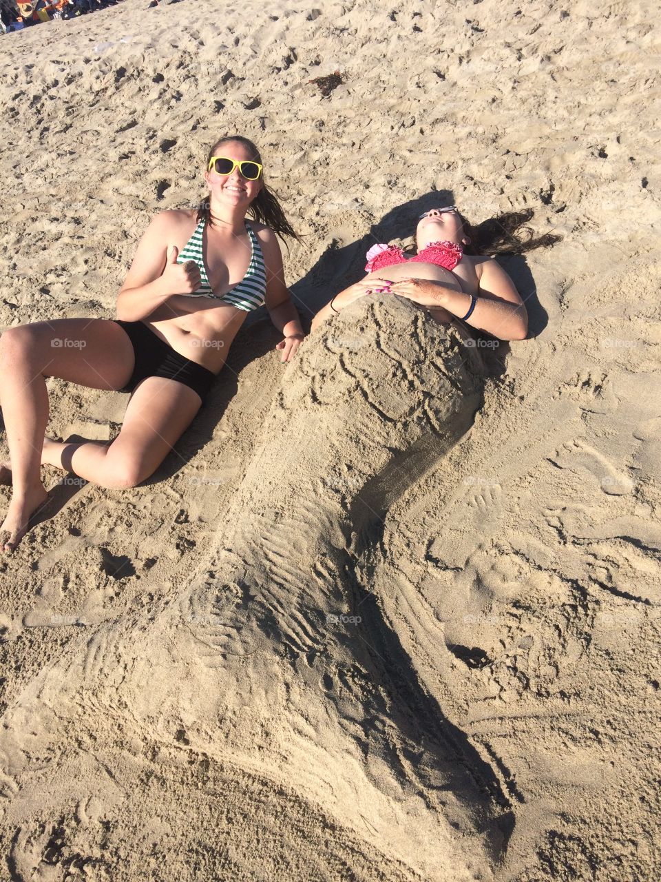 Two girls relaxing on a California beach in October, one enveloped in sand to look like a mermaid tail.
