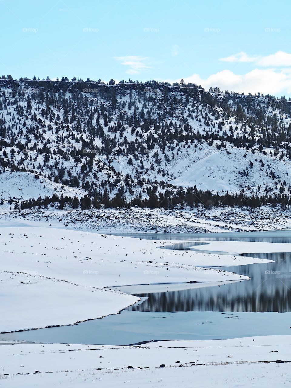 Hills covered in fresh snow and bright skies reflecting on the waters at Juniper Point at Prineville Reservoir in Central Oregon on a winter day.                                                    