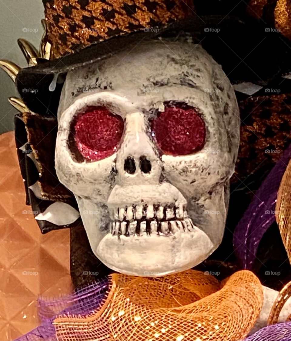 A close-up view of a human skull with red eyes wearing a black hat and surrounded by orange sparkly fabric