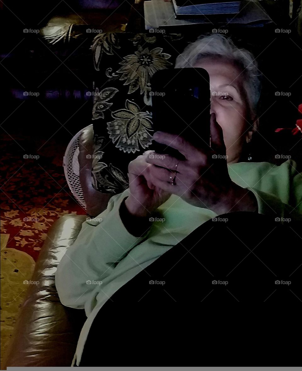 Girl laying on couch working oncell phone in dim light. She's intently involved in what's she is texting or reading. Her expression doesn't seem worried because of the pleasant look on her face.