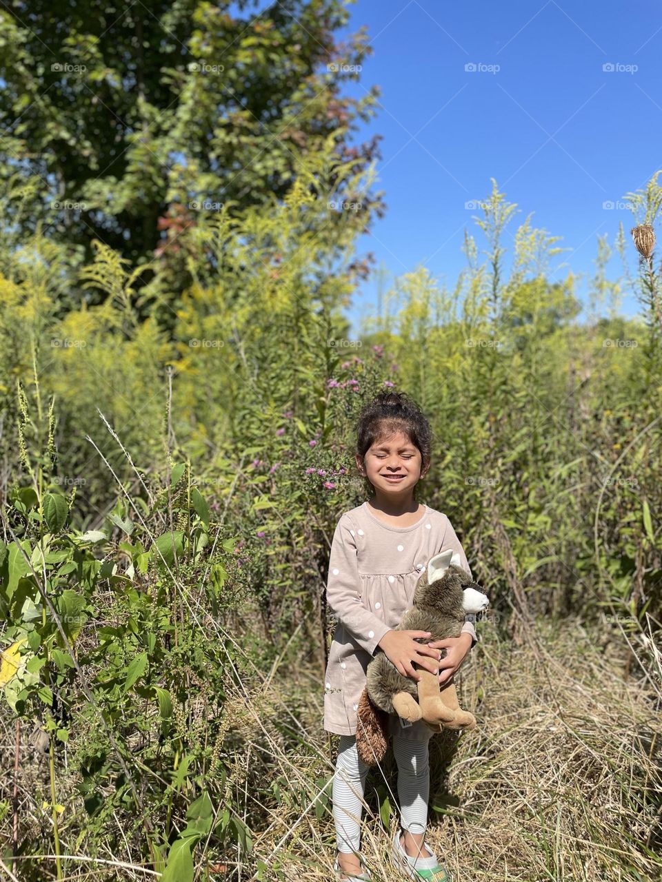 Little girl poses with coyote in the woods, too sunny for toddler’s eyes, little girl squinting into the sun, posing with stuffed animals in the park