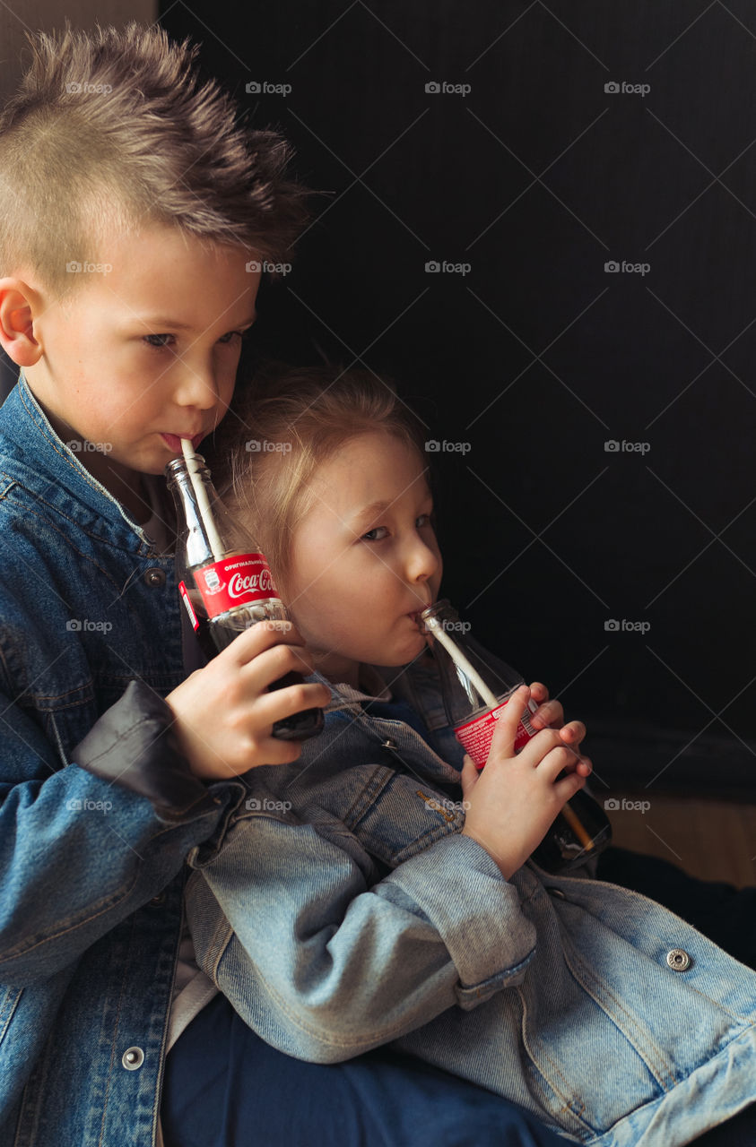 brother and sesira, stay at home, dressed in denim clothes, drink Coca-Cola from bottles through a straw.  beautiful stylish kids