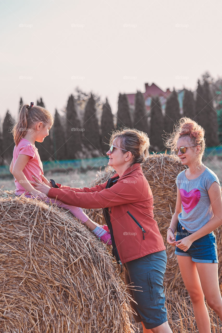 Mother and her daughters, teenage girl and her younger sister playing together on hay bale outdoors, spending vacations in the countryside. Candid people, real moments, authentic situations