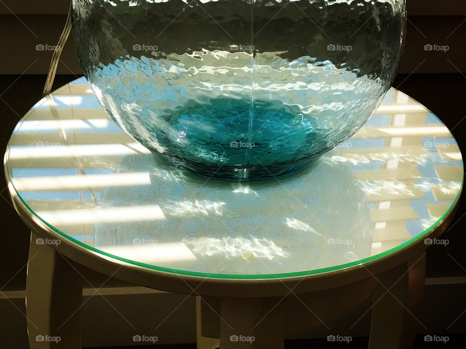 The sun was shining through the rectangular wooden blinds producing rectangular reflections of the blinds & the blue sky on the glass topped table; even through the transparent, textured, glass lamp. 