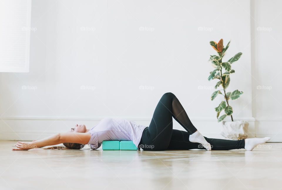 Young woman in sports uniform doing stretching on fitness blocks lying on floor in bright studio