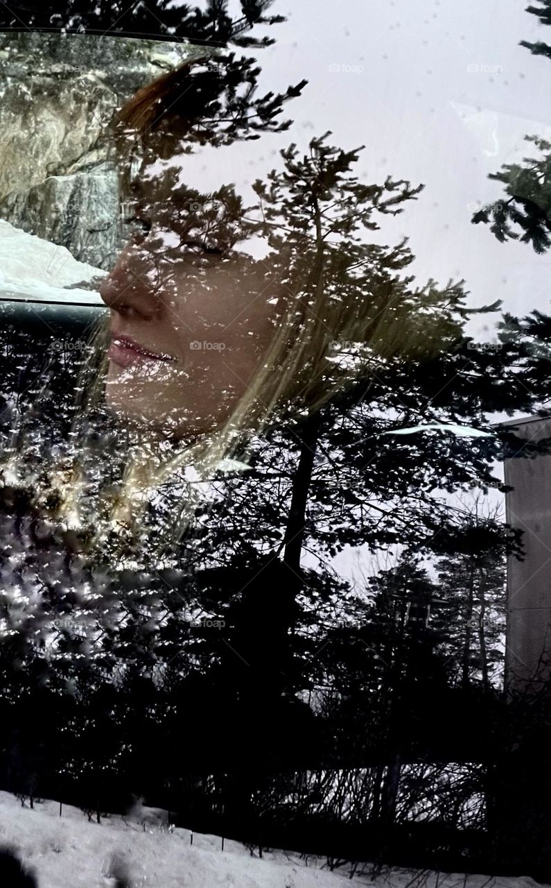 A woman looks out the car window. The reflection of the trees on window connects to the face.