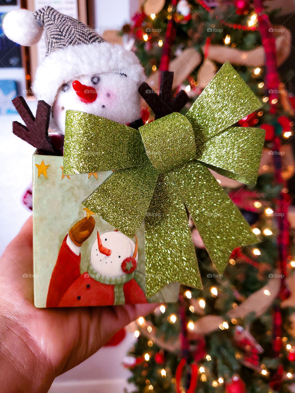 Bright plush mini snowman in a snowman box with lighted Christmas tree in the background.