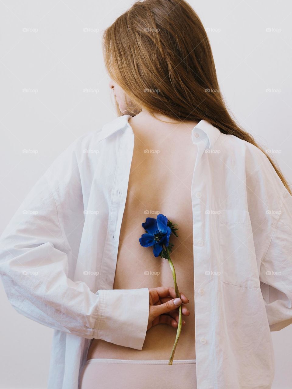 Young beautiful girl with long red hair wearing white shirt holding blue anemone on white background, portrait of woman 