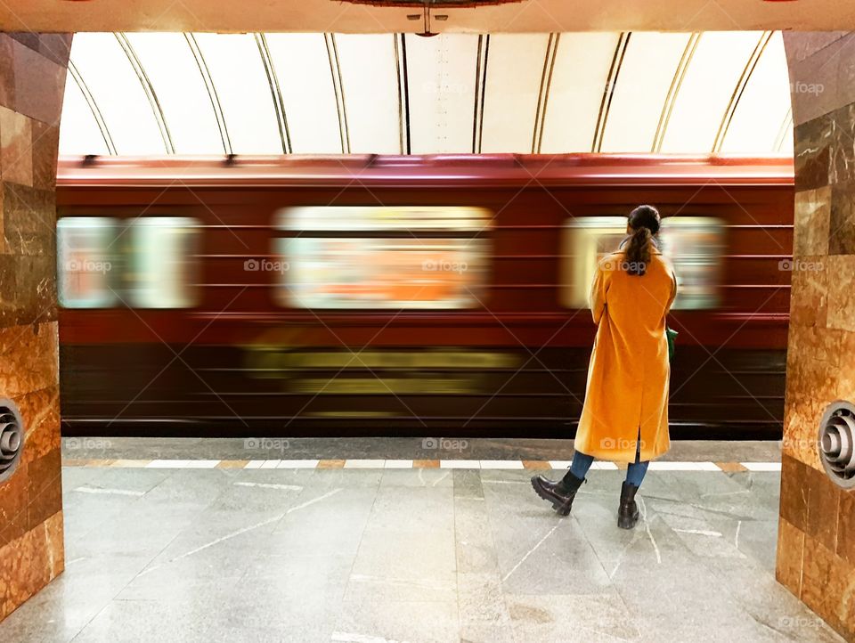 Young woman in yellow coat standing in front of a moving subway train 