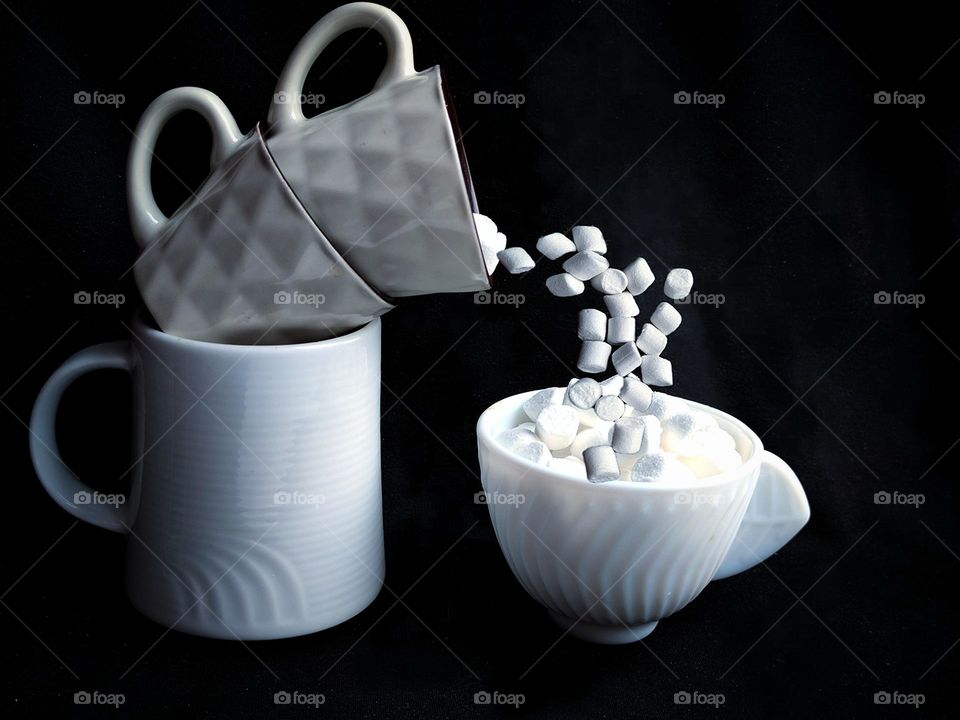 White colour. The background is black. Three white glasses stand on top of each other in the form of an arc. White marshmallow is crumble from the top glass into a white cup with white marshmallow