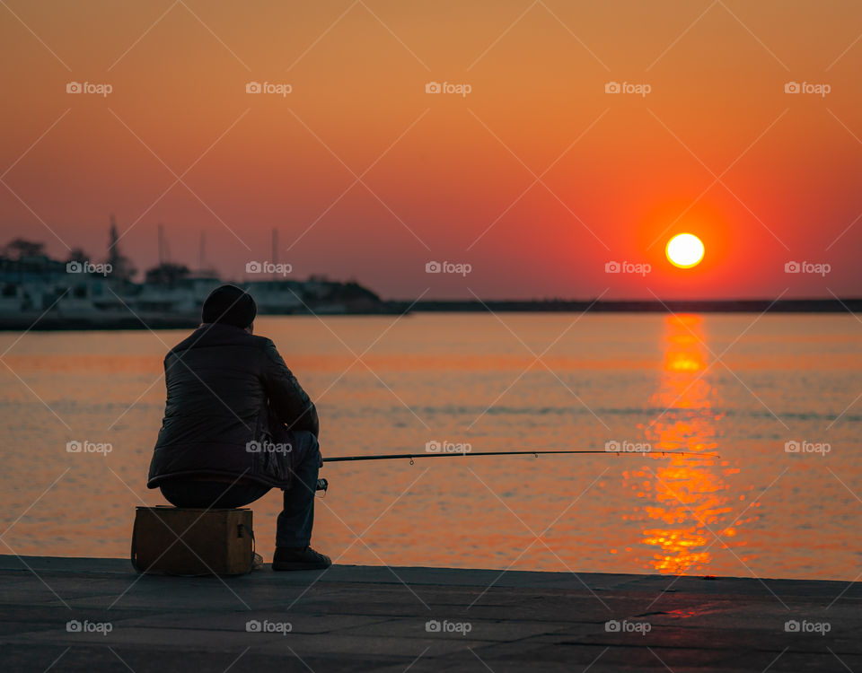 A fisherman sits by the sea on a pier during sunset and catches a fish.