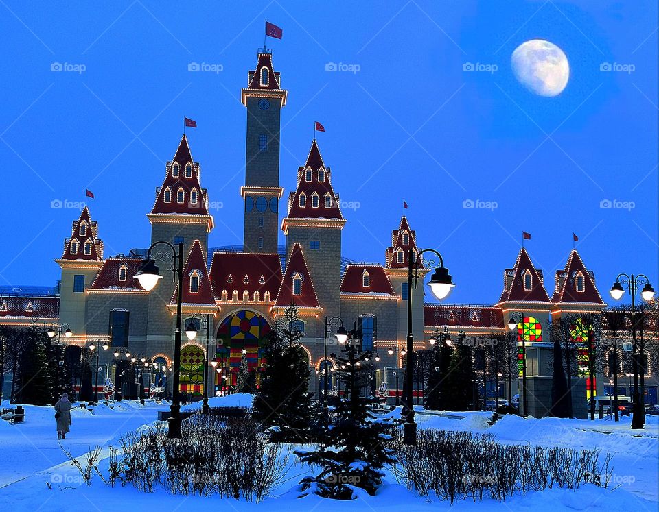 Winter.  Amusement complex "Dream Island" made of stone, with towers, decorated with a luminous garland.  Twilight.  Above the complex is a large moon.  In the foreground trees and firs in the snow