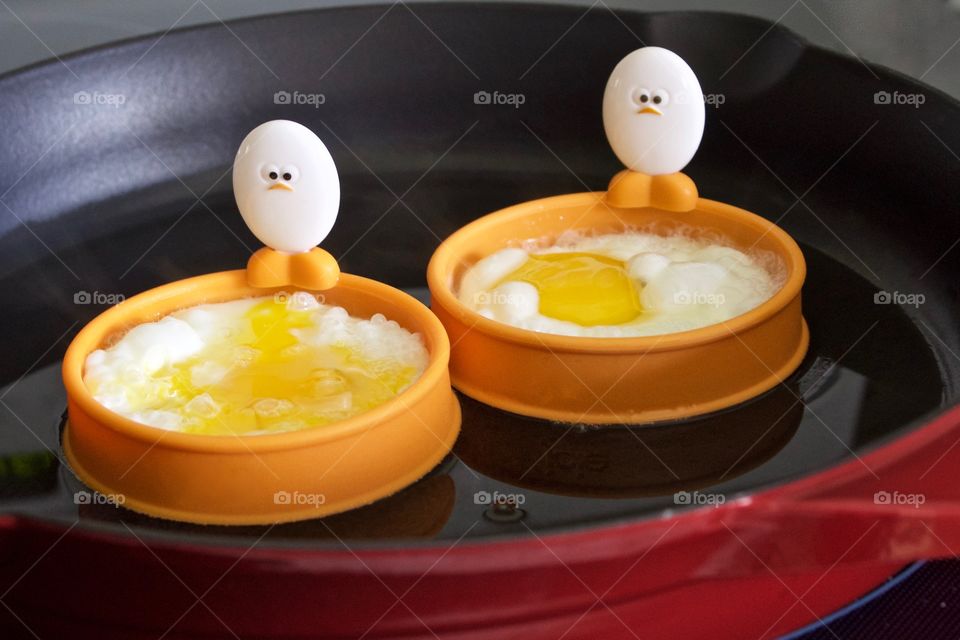 Closeup of a cast iron pan containing eggs being fried in silicone egg rings with egg-shaped handles depicting baby chick faces and feet