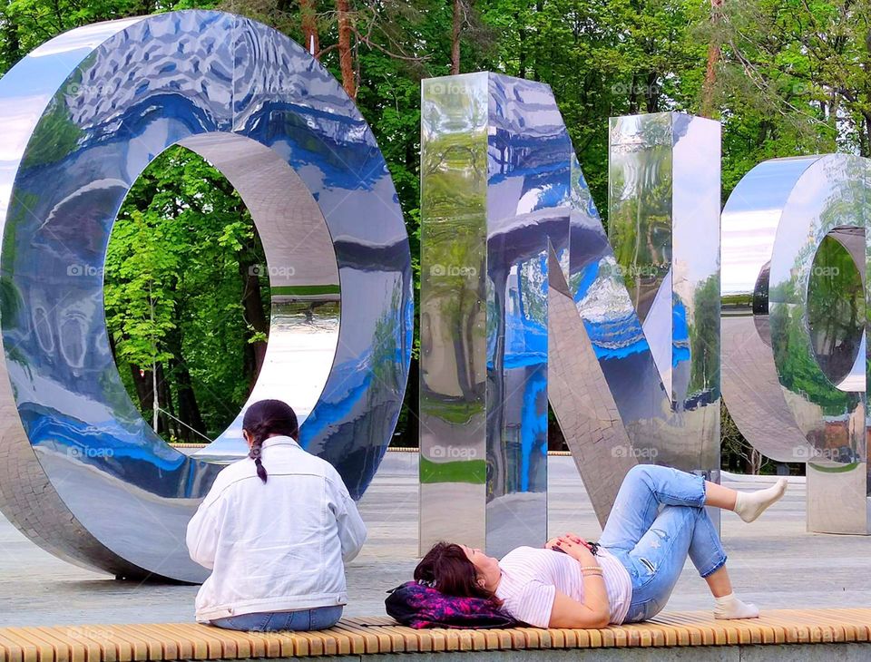 Summer in the city.  There are two women on the bench.  One woman lies and rests