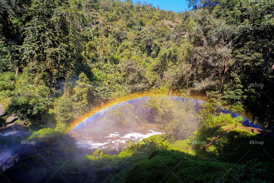 This is the suburb of Chiang Mai, Thailand. I stand next to the waterfall and look down. A beautiful rainbow connects the forest and the bridge. It is a perfect moment!
