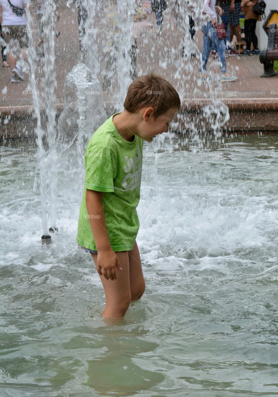 child boy in the water splash fountain, summer heat, city street view
