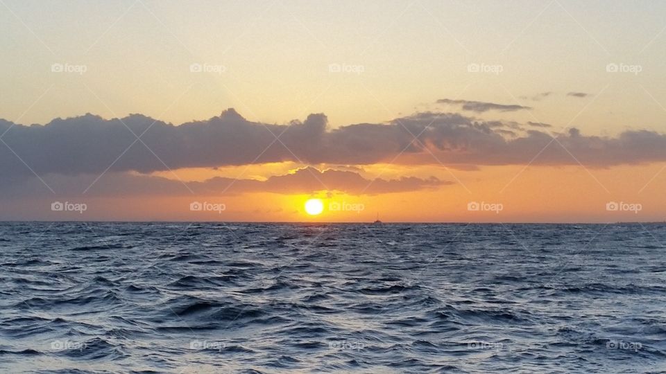 View from ocean of beautiful sunset. Colors of blue, orange, yellow, and grey. Waves of the sea, and sun in clouds, in Kauai, Hawaii.