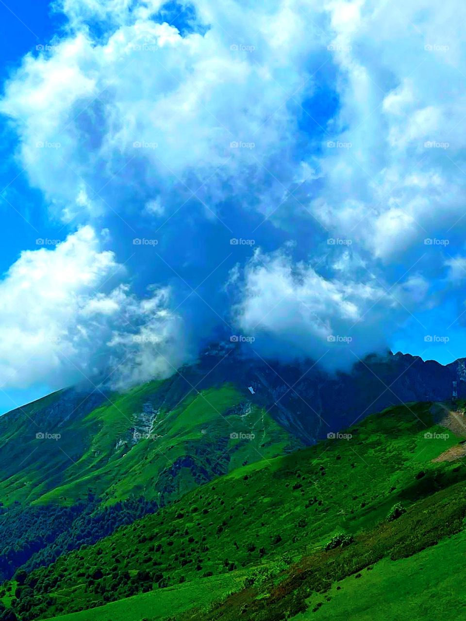 Summer.  Green mountains, the peaks of which are covered by clouds against the blue sky