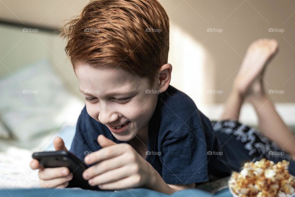 Red-haired boy watching a movie on the phone lies on the bed