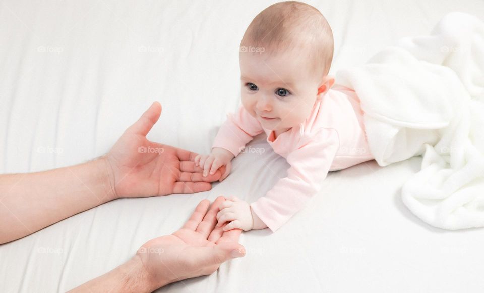 A beautiful caucasian baby stretches his arms to his father's hands while lying on a bed with white linens, close-up from the side. The concept of newborns, happy childhood, home sweet home.