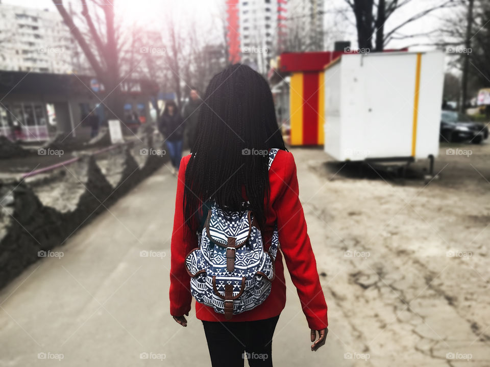 Young woman wearing a red coat and a blue backpack walking through the city 