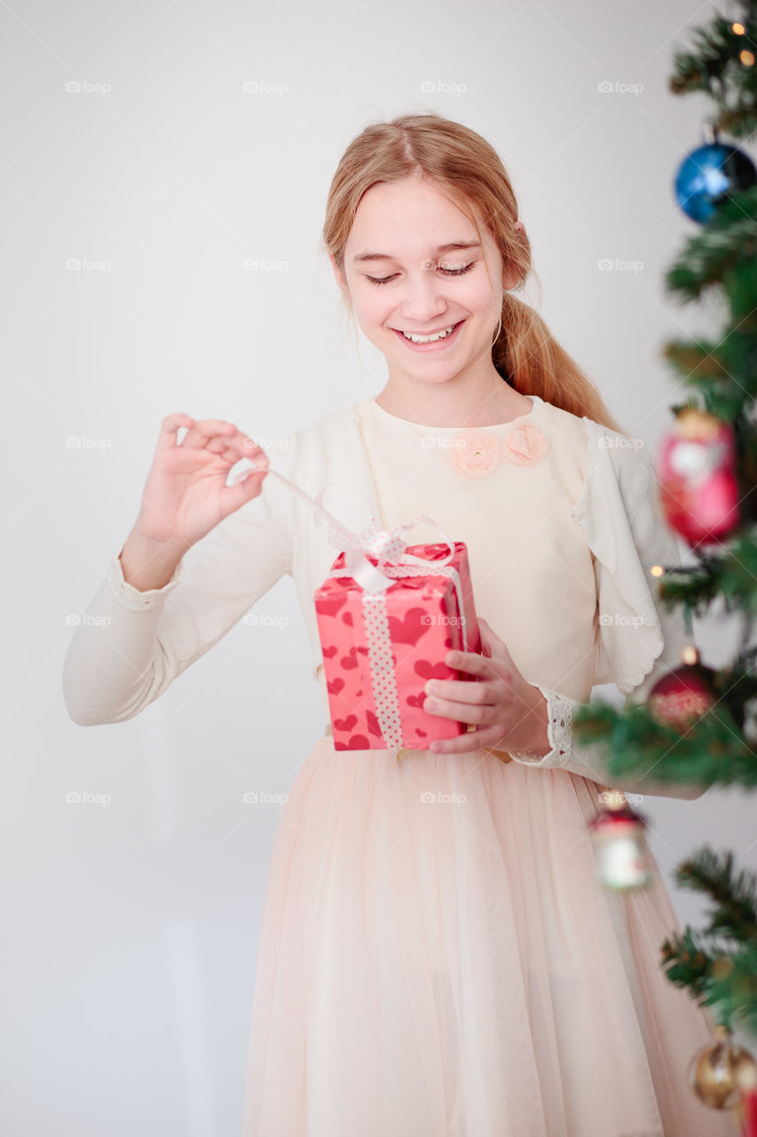Happy smiling girl wearing pink dress standing behind a tree and  unpacking Christmas gift