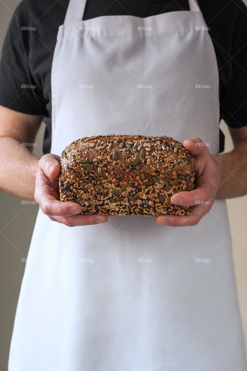 Close-up at bakers hands holding a multi-seeded loaf of whole grain bread in front of him.