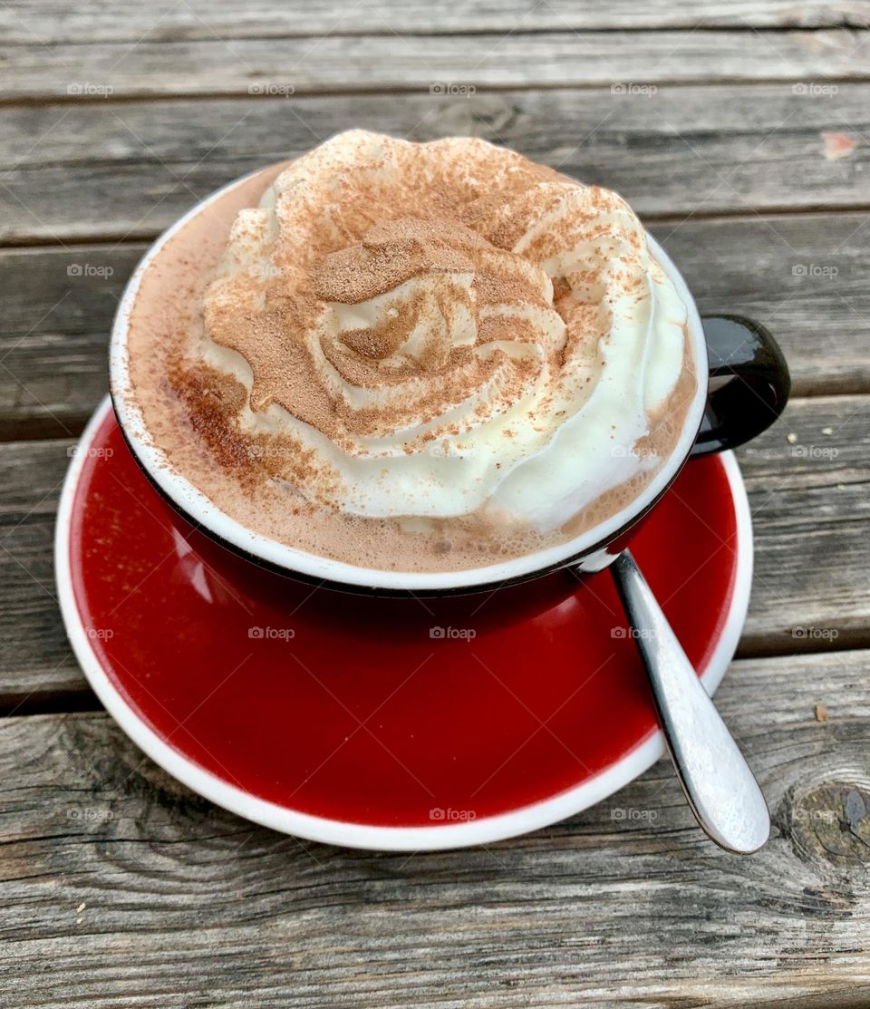 Cup of hot chocolate in a dark cup with deep red saucer and a metal teaspoon is on a wooden table