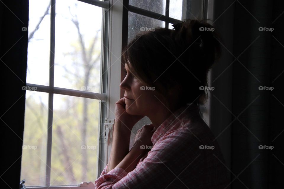 Sad Woman looking out window wearing pink long sleeve shirt yellow leaves on tree outside window