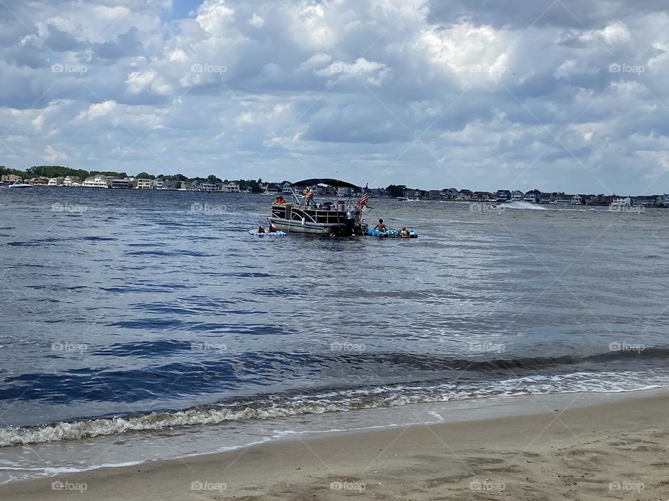 People boating and rafting in Silver Bay viewed from the beach in Cattus Island Park in Toms River, NJ. Waves lap gently against the shoreline. Puffy clouds fill the sky, and the water is blue and beautiful. It’s a great place to relax and have fun. 
