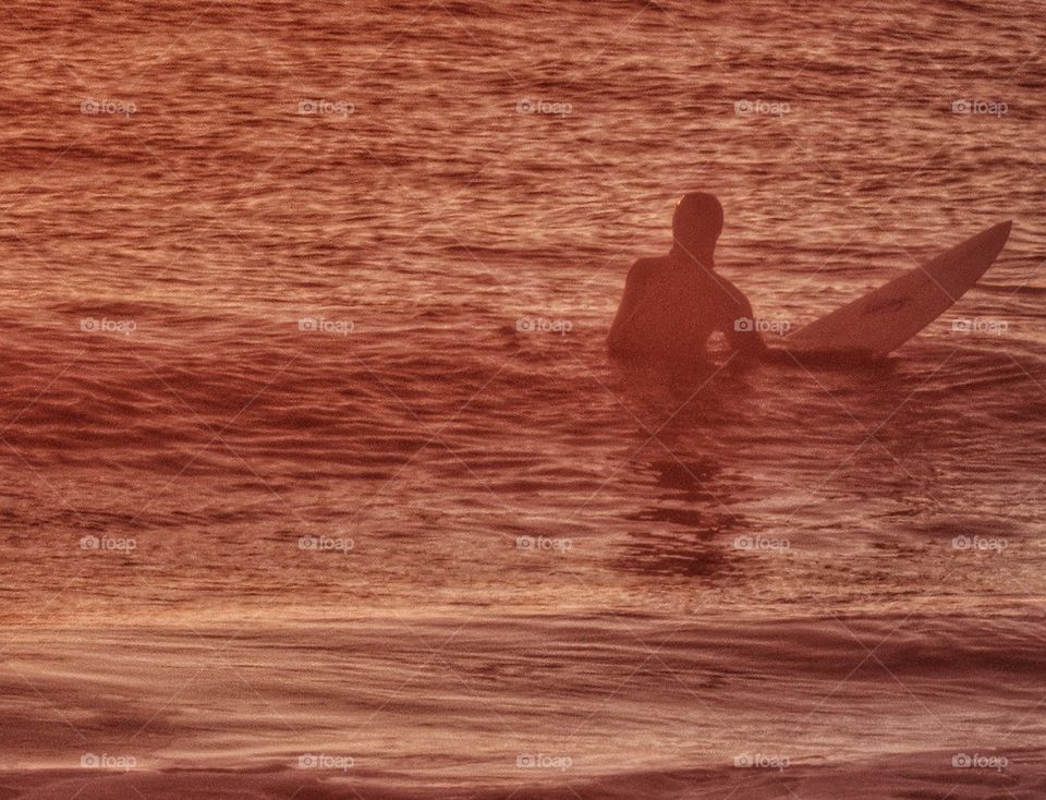 California Surfer At Sunset. Surfing In Water Turned Red By The Sunset