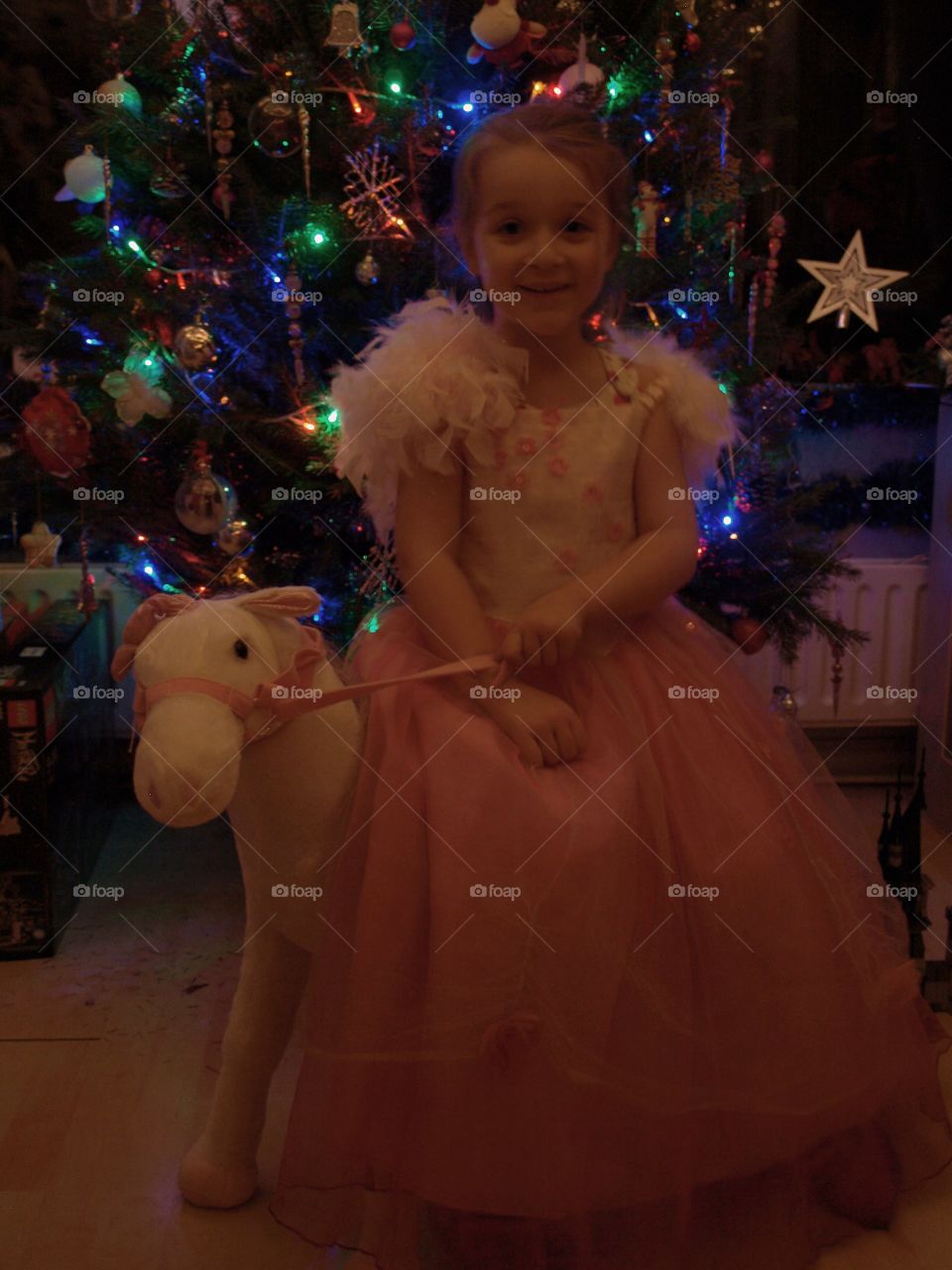 little girl dressed in a beautiful pink formal dress on a unicorn. in front of a lit Christmas tree. Christmas eve.