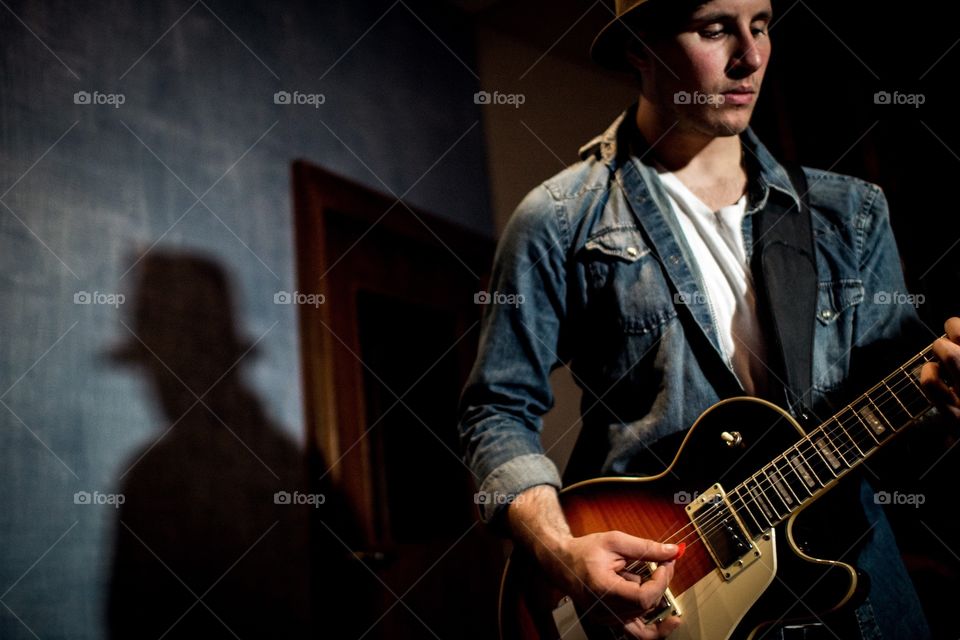 Guy wearing hat playing guitar in a recording studio. Rock and roll guitar player wearing denim with blue walls