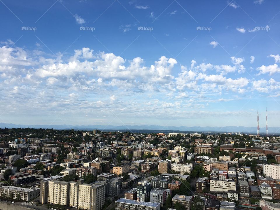 Downtown buildings viewed from a rooftop apartment. The sky is clear blue with some white clouds. The skyline buildings can be seen from the top. Mountain valleys in the distance. 