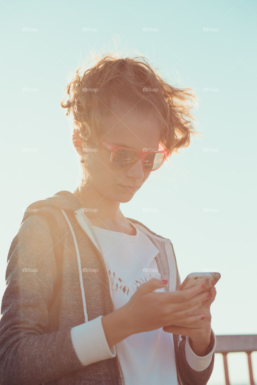 Young blond hair girl using phone, looking at screen, standing outdoors, she is backlighted by sunlight with plain sky in the background