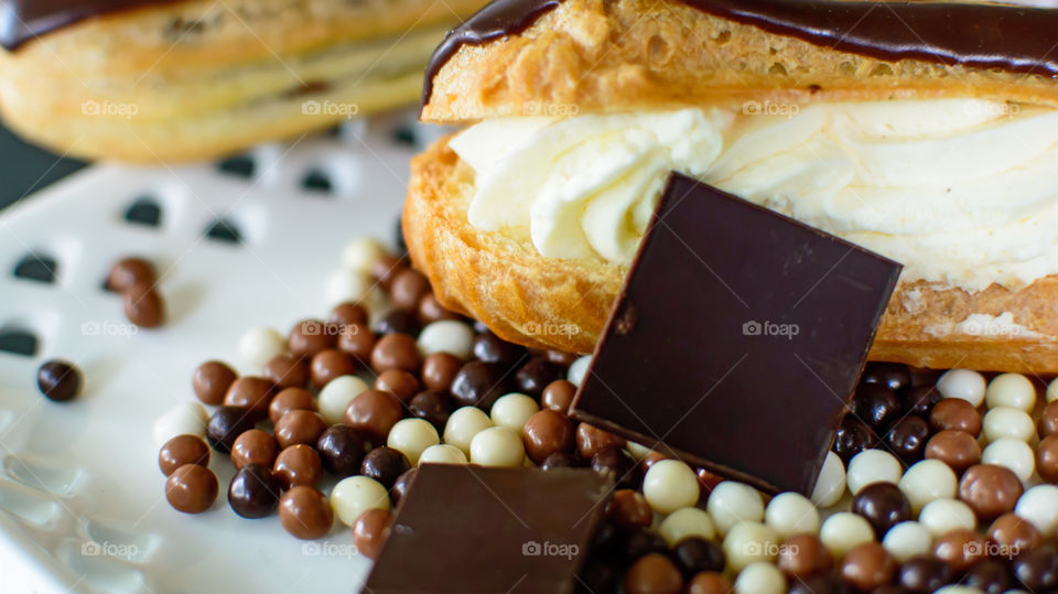 Chocolate ganache coated cream puff pastry eclair filled with whipped cream with 80% dark chocolate squares and white chocolate, milk chocolate and dark chocolate balls on plate ready to eat dessert