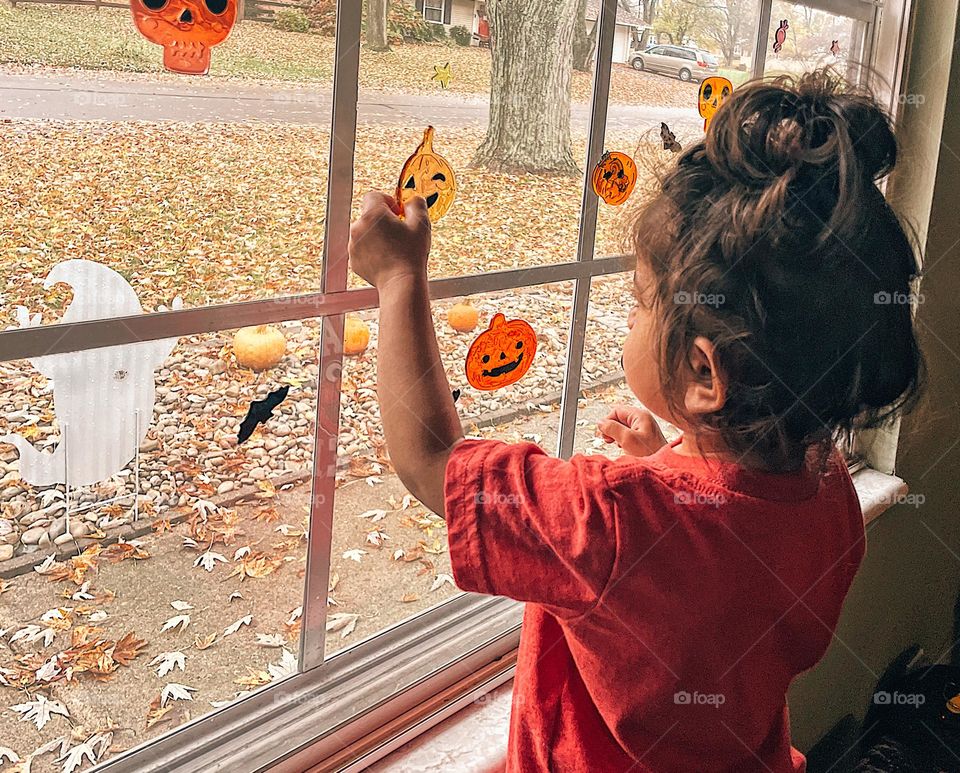 Child putting up Halloween decorations, child placing Halloween clings on window, toddler playing with Halloween decorations on window, toddler enjoying Halloween decorations, Halloween window clings, spooky decor with toddlers