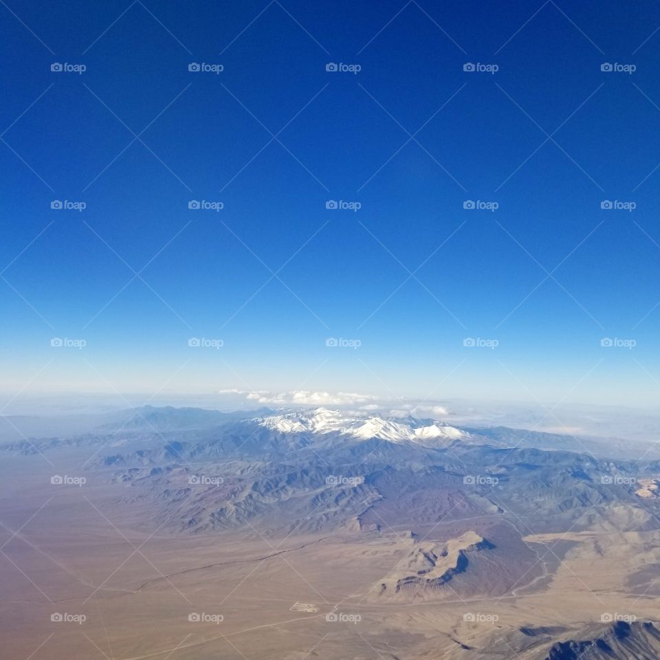 airplane view of dry desert terrain and snow covered peaks with blue sky