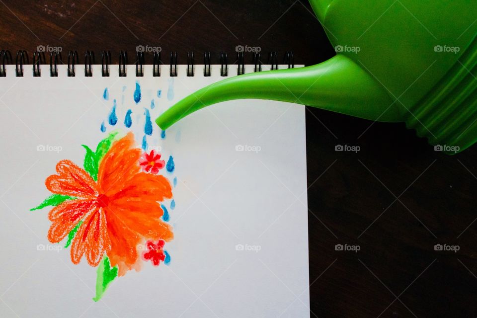 Flat lay watercolor crayon drawing in a mixed media notebook of a large orange flower being “watered” by a green watering can on a dark wood surface