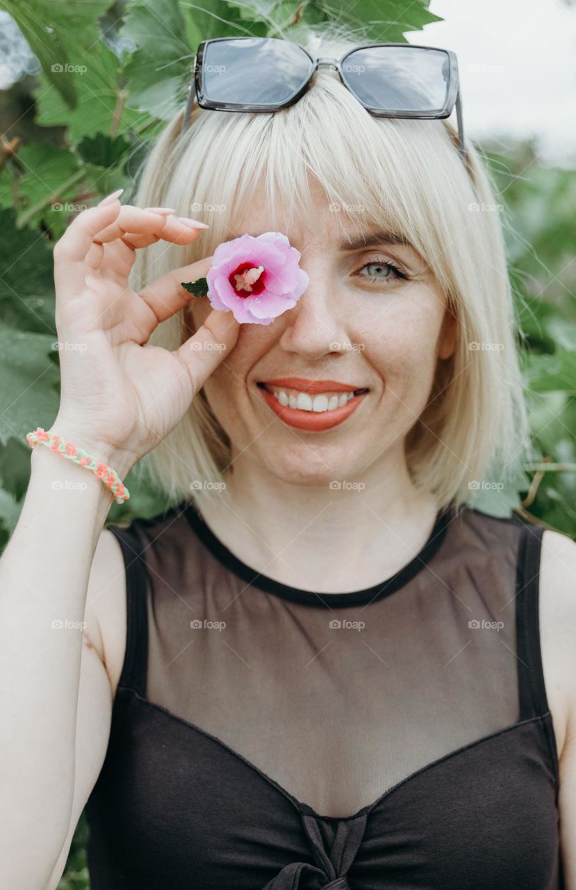 Portrait of one beautiful Caucasian happy blonde girl with a smile holding a pink flower with one hand on her eye, looking at the camera and standing against the background of a bush on a summer day, side view close-up.