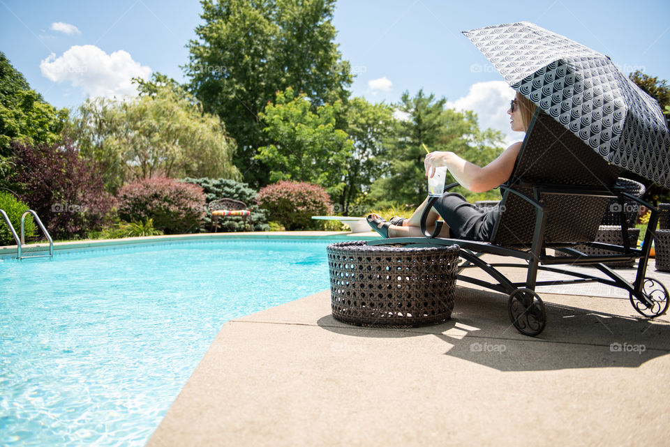 From behind view of a woman sitting poolside in a lounge chair and holding an umbrella
