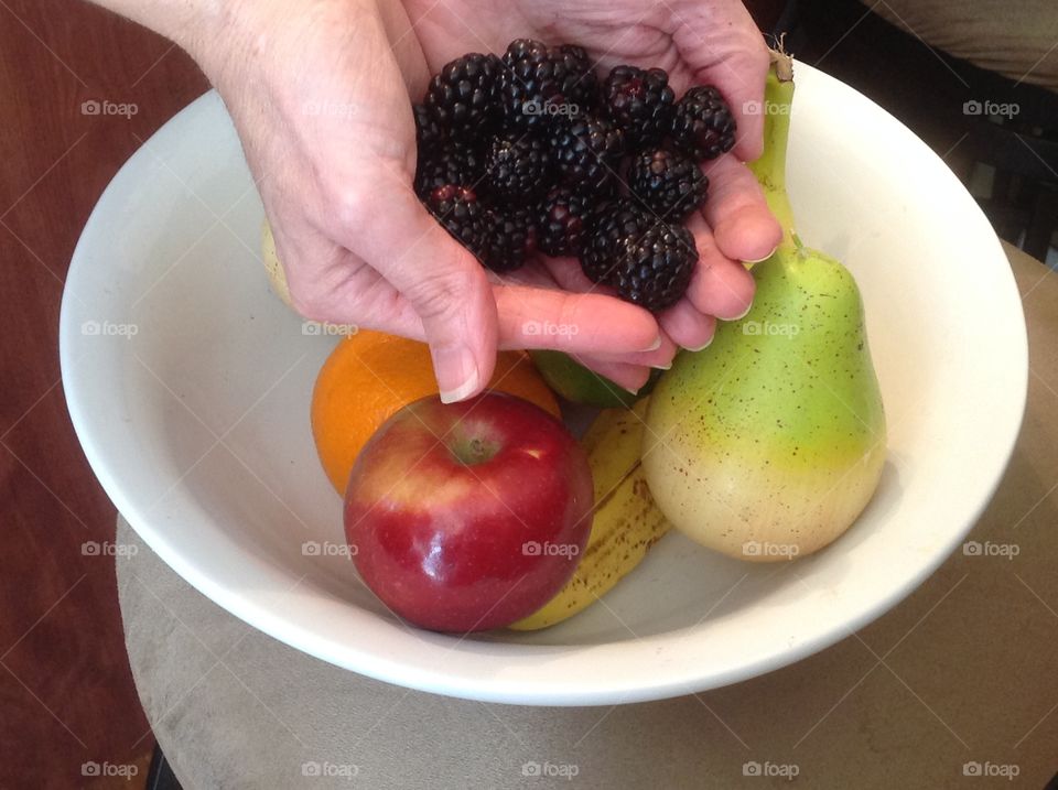 Holding in hand blackberries near a bowl of colorful fruit.