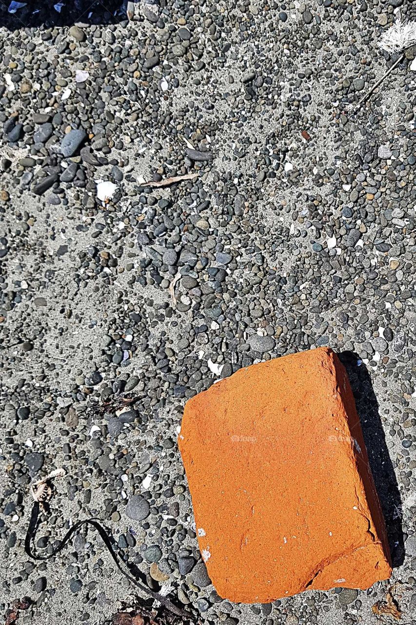 A piece of orange brick lays rested in the sea shore sand. Stands out against the grey background of stone and sand.