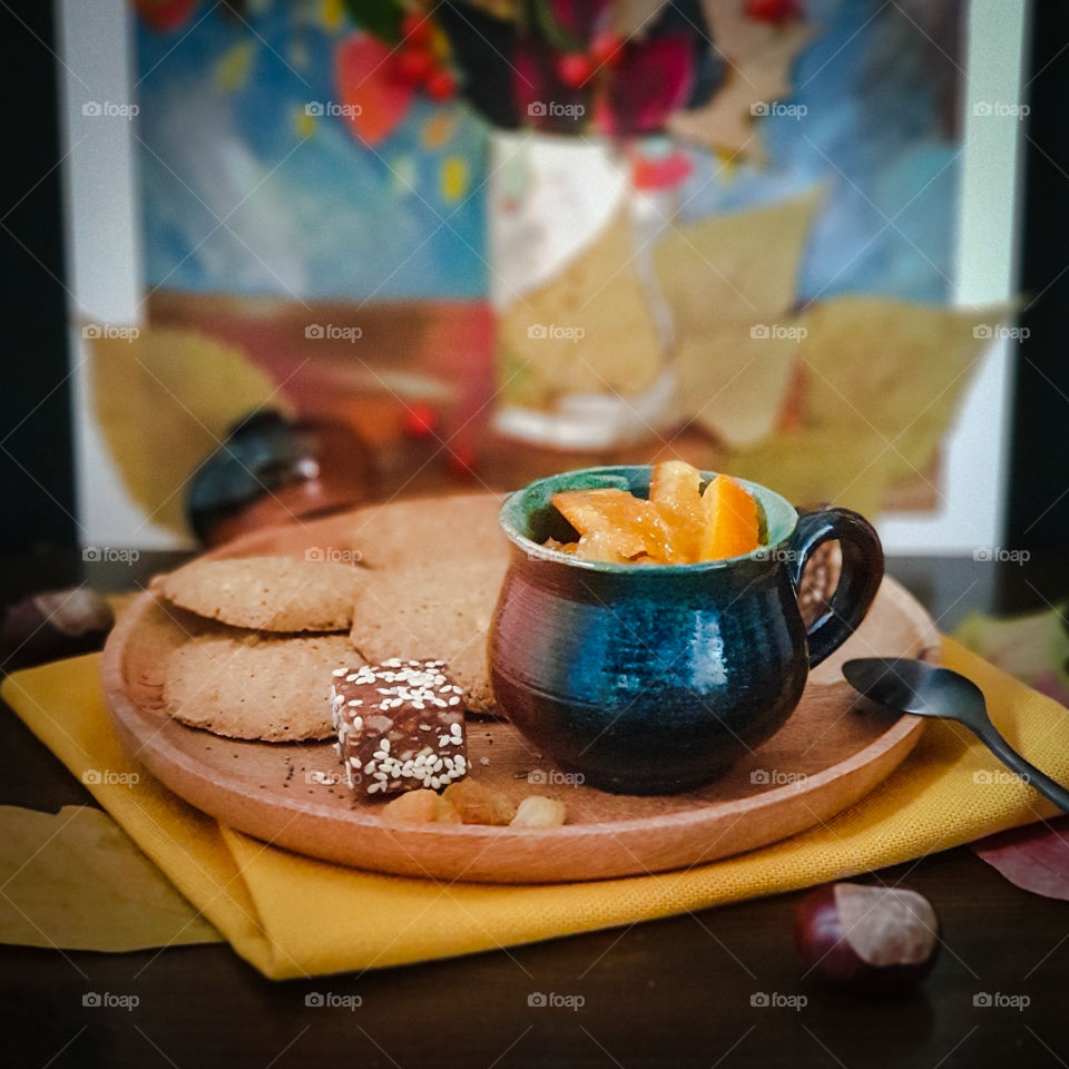 Autumnal picturesque still life.  Wooden tray with clay pot with orange jam and crumbly cookies, chestnuts and sweets on a bright yellow napkin on the background of a child’s applique picture with autumn leaves and mountain ash