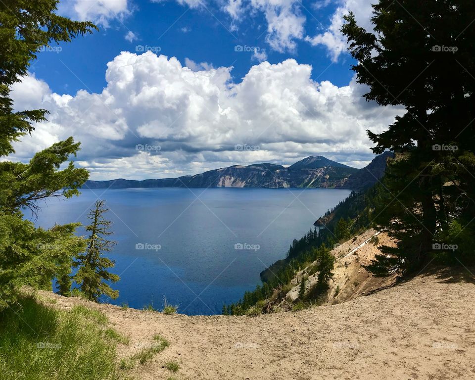 Vibrant Landscape photography of Crater Lake at Crater Lake National Park in Oregon,USA; Bright cloudy sky in background, with vibrant blue lake water, and green and dark trees in foreground
