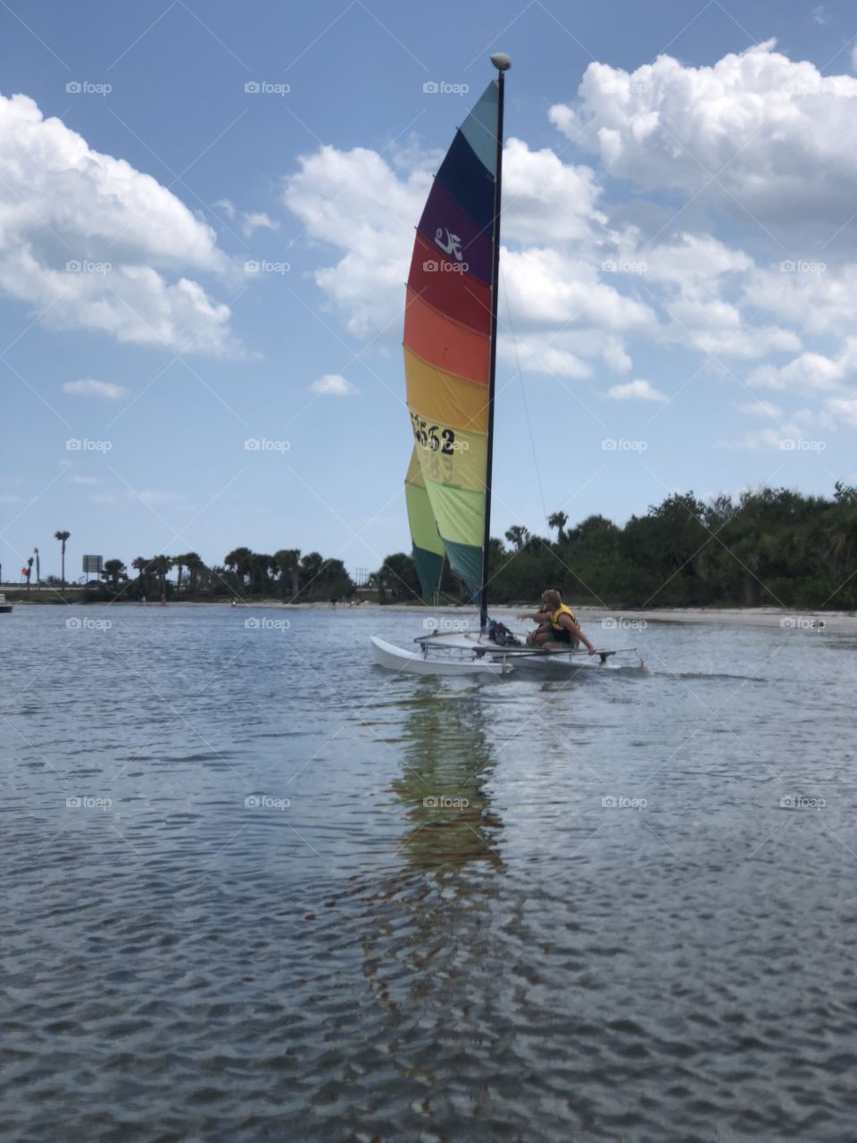 Colorful sail boat with man on it in the river , trees and clouds in background 