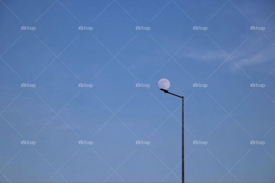 The full moon in the clear blue sky above a street lamp during the day 