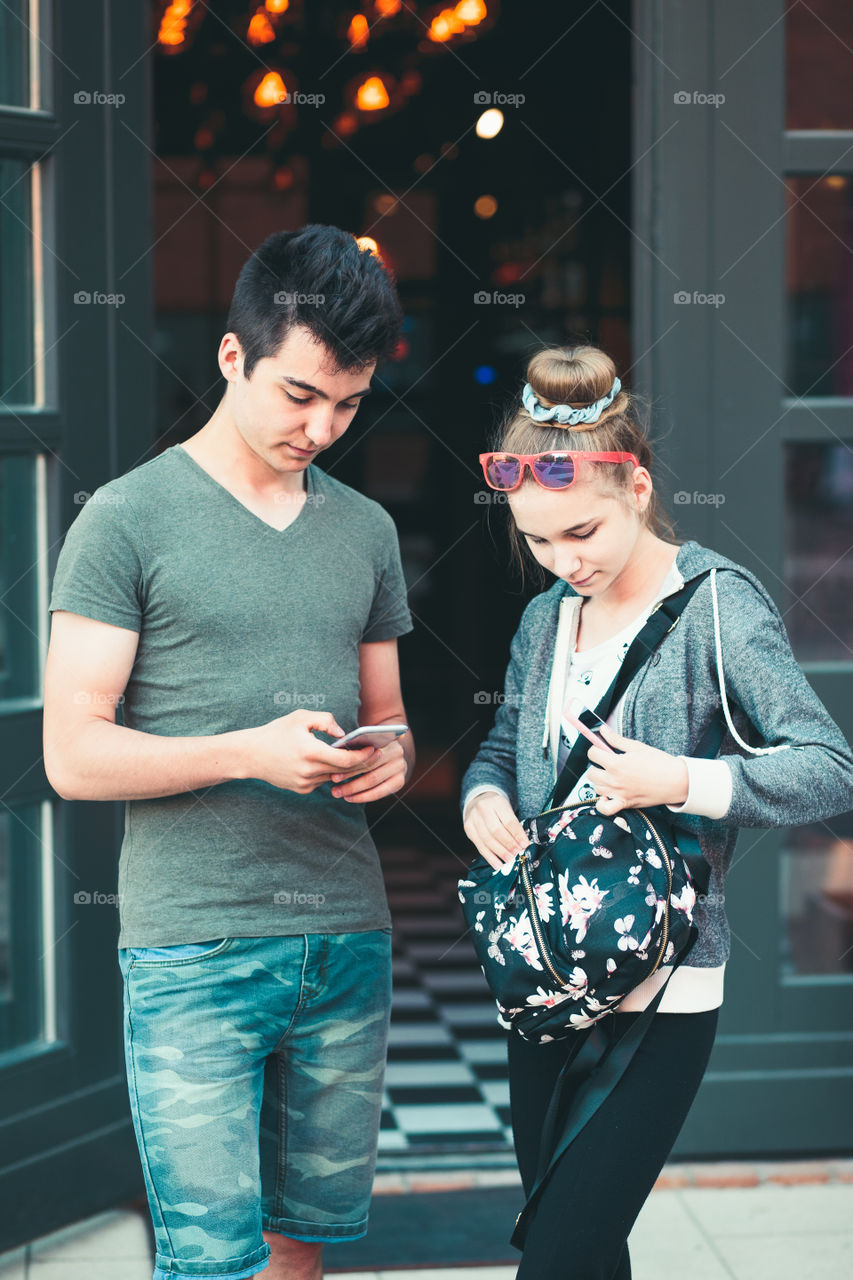 Couple of friends, teenage girl and boy, using smartphones, talking together, standing on street in center of town, spending time together