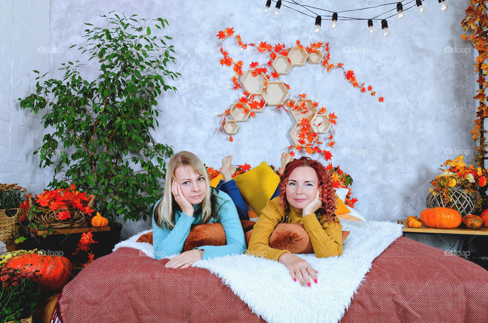 Two lovely women, sisters, friends in warm sweaters sitting in the bed at a cozy home, laughing, enjoying. Home interior with autumn decorations, orange leaves, flowers, pumpkins.