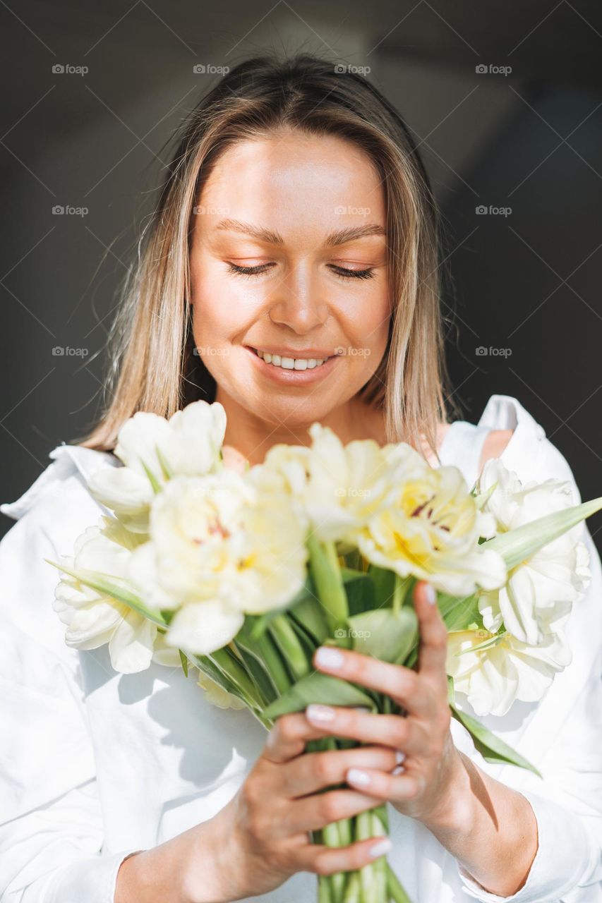 Young beautiful smiling woman forty year with blonde long hair in white shirt with bouquet of yellow flowers in hands near window in bright interior at the home